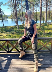 Kvinde i stille skovbukser sidder paa traebro med soe og skov i baggrunden