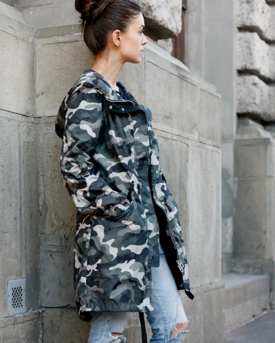 Pige klaedt i sneulv camouflage jakke foran gammel bygning der kigger væk